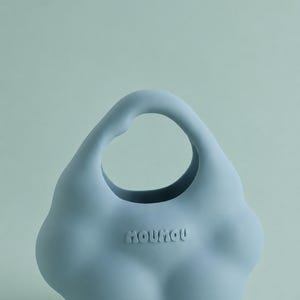 Puede incluir: Un bolso de mano de silicona de color azul claro con forma de nube y un asa circular integrada. El bolso presenta un acabado liso y mate con la palabra MOUMOU grabada en una tipografía redondeada y burbujeante en el centro frontal.