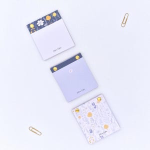 May include: Three floral notepads with gold accents and a gold paperclip on a white background. The notepads feature floral patterns in blue, yellow, and white. Each notepad has the name "Melissa Sophia" printed on it.