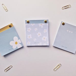 May include: Three pale blue notepads with gold detailing and floral patterns, secured with gold studs. Gold paper clips are scattered around the notepads on a white surface.