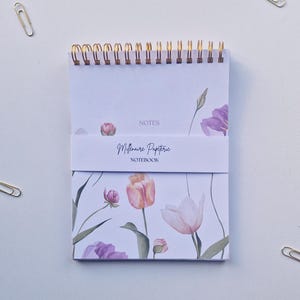 May include: A white spiral notebook with a floral design featuring pink and white tulips and green leaves. The notebook has a gold wire binding and a label that reads "Millenne Paperie Notebook".