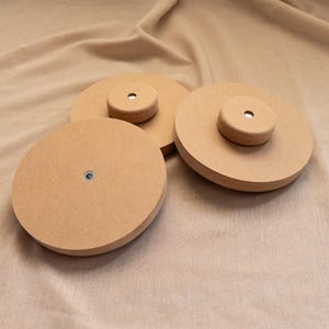 Könnte beinhalten: Drei kreisförmige, hellbraune Holzscheiben in verschiedenen Größen, die jeweils ein kleines, erhöhtes zylindrisches Mittelstück mit einer sichtbaren Metallbefestigung oder einem Loch in der Mitte aufweisen, angeordnet auf einem neutralen Stoff.