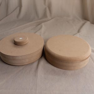 Könnte beinhalten: Zwei kreisförmige, hellbraune Holzbehälter mit glatten, flachen Oberflächen. Ein Behälter verfügt über einen kleinen, mittig platzierten runden Knopf auf dem Deckel, während der andere einen schlichten, flachen Deckel hat. Einfaches, geschichtetes Design.