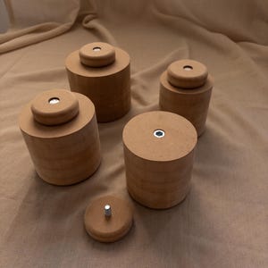 Könnte beinhalten: Ein Set aus vier zylindrischen, hellbraunen Holzmöbelfüßen, die jeweils eine flache Oberseite mit einer mittigen Metallbolzenbefestigung aufweisen. Eine zusätzliche kleine, runde Holzscheibe mit einer hervorstehenden Metallschraube liegt davor.