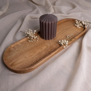 Könnte beinhalten: Ein ovales Serviertablett aus Holz mit einer dunkelgrauen Kerze und weißen Trockenblumen auf einem weißen Stoffhintergrund.