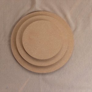 Könnte beinhalten: Drei kreisförmige, flache Holzplatten in verschiedenen Größen, die übereinander gestapelt sind. Die Platten bestehen aus einem hellbraunen, gepressten Holzfasermaterial mit einer glatten, gleichmäßigen Oberfläche und sichtbarer Maserung.