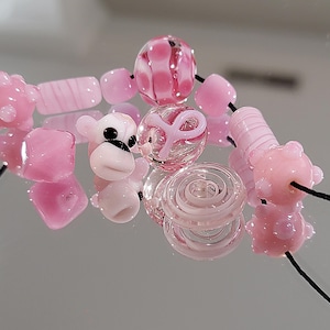 May include: A collection of pink glass beads in various shapes, including round, square, and cylindrical. Some beads have internal designs, and one features a monkey face. The beads are strung on black cord, reflecting on a mirrored surface.