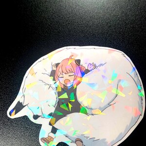 May include: A sticker featuring a cartoon girl with pink hair and a black dress, sleeping on a large white furry animal with iridescent glitter.