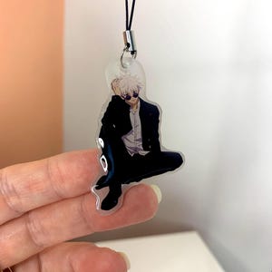 May include: A clear acrylic keychain featuring a cartoon character with white hair, wearing a black jacket, black pants, and sunglasses. The keychain is attached to a black string.