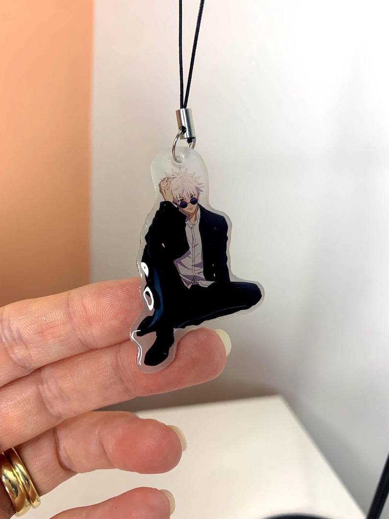 May include: A clear acrylic keychain featuring a cartoon character with white hair, wearing a black jacket, black pants, and sunglasses. The keychain is attached to a black string.