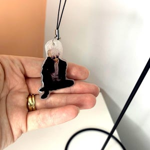 May include: A clear acrylic keychain featuring a black and white illustration of a person with white hair wearing sunglasses and a black suit. The keychain is attached to a black string.