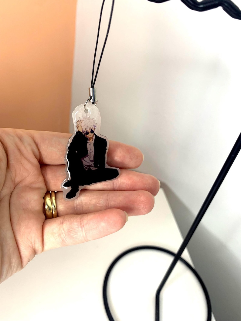 May include: A clear acrylic keychain featuring a black and white illustration of a person with white hair wearing sunglasses and a black suit. The keychain is attached to a black string.