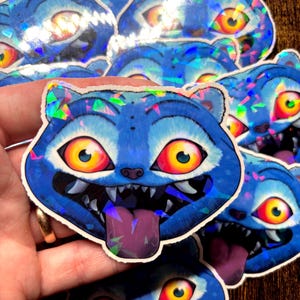 May include: A selection of blue cat-shaped stickers with a holographic effect. Each sticker depicts a cat face with large yellow and red eyes, sharp teeth, and a purple tongue. The stickers have a white border.