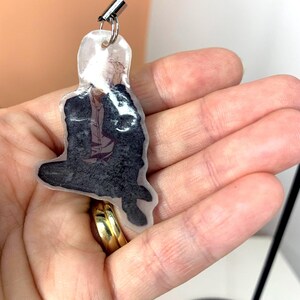 May include: A clear plastic keychain with a black and white image of a person sitting down. The keychain has a black string attached to it.