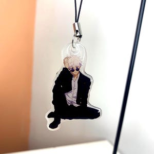 May include: A clear acrylic keychain featuring an anime character with white hair, wearing a black suit and sunglasses. The character is sitting with one leg crossed over the other.