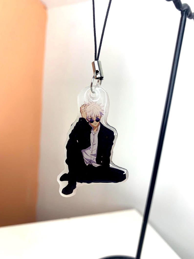 May include: A clear acrylic keychain featuring an anime character with white hair, wearing a black suit and sunglasses. The character is sitting with one leg crossed over the other.