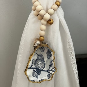 Può includere: Un fermatenda decorativo con un guscio di ostrica dipinto con un disegno di uccello e fiori. Il guscio è rifinito con dettagli dorati ed è attaccato a una fila di perline di legno color crema con dettagli dorati.