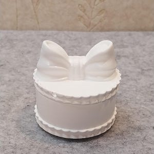 Könnte beinhalten: Kleine, weiße, runde Schmuckschatulle mit einem Deckel in Schleifenform. Die Schatulle hat ein gewelltes Randdesign sowohl auf dem Deckel als auch auf dem Boden, was einen dekorativen Touch verleiht. Hergestellt aus glänzendem Material.