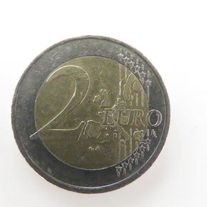 Puede incluir: Una moneda de dos euros de plata con un centro dorado que muestra el número "2" y la palabra "EURO". La moneda tiene un diseño de un edificio con una torre y estrellas alrededor del borde.
