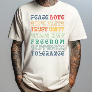 Può includere: Una maglietta bianca con una stampa grafica colorata delle parole "Peace Love Hope Faith Trust Unity Harmony Freedom Happiness Tolerance".