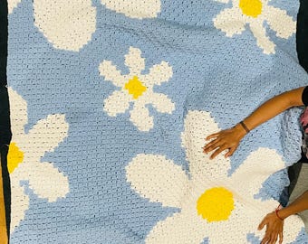 May include: A large, handmade crochet blanket featuring a daisy pattern. The blanket has a light blue background with large white daisy petals and yellow centers. The blanket is made of soft yarn and is perfect for a bed or couch.