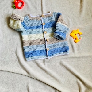 May include: A handmade, striped crochet baby cardigan in shades of blue, white, and tan, with wooden buttons. The baby sweater is displayed with two colorful baby toys. The cardigan is a cozy, handmade item.