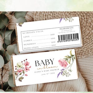 May include: Two rectangular candy bar wrappers with floral designs and text. One wrapper displays "Nutrition Facts" and "Ingredients." The other wrapper reads "BABY in bloom" and "Avery's Baby Shower June 12th 2025." The wrappers are on a textured, neutral-toned surface.
