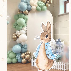 May include: A Peter Rabbit party decoration with a large cutout of the character wearing a blue jacket. The cutout is in front of a white arch and a balloon garland in shades of blue, green, gold, and white. A small white picket fence is in front.