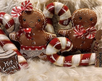 May include: Two gingerbread people ornaments with red and white candy cane accents, a red and white candy cane, and a brown mug with the words "Hot Cocoa" on it. The ornaments are on a white fluffy surface.