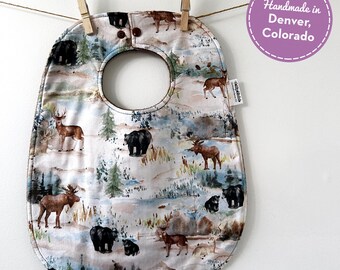 May include: A baby bib with a forest animal print, including deer, bears, and moose, hangs from a clothesline. The bib has a large neck opening and is secured with two wooden clothespins. The text "Handmade in Denver, Colorado" is in a purple circle.