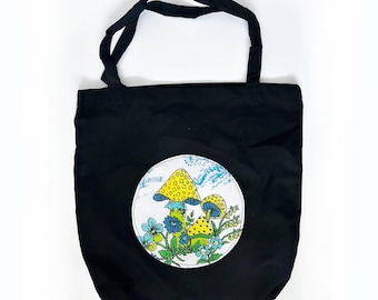 May include: Black canvas tote bag with a white circle design featuring three yellow mushrooms with blue and green accents. The mushrooms are surrounded by blue flowers and green leaves.