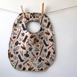 May include: A beige baby bib with a black trim and a pattern of equestrian-themed illustrations, including horseshoes, saddles, boots, and riding helmets.