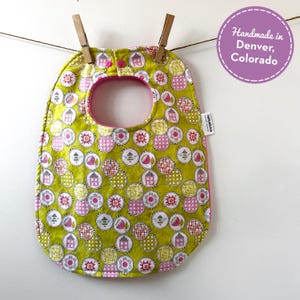 May include: A handmade baby bib with a green background and a pattern of pink, white, and orange circles. The bib has a pink trim and a button closure. The bib is hanging from a clothesline with two wooden clothespins. The text "Handmade in Denver, Colorado" is in a purple circle.