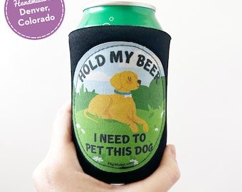 May include: Black beer can cooler with a graphic of a yellow dog wearing a blue collar, with the text "HOLD MY BEER, I NEED TO PET THIS DOG." The cooler is holding a green can. The text "Handmade in Denver, Colorado" is in a purple circle.