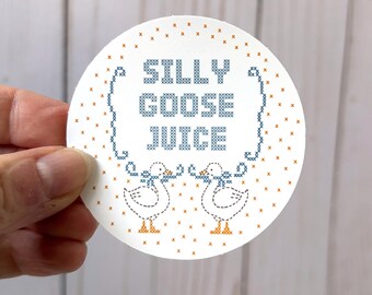 May include: A circular white sticker featuring a cross-stitch style design. The text SILLY GOOSE JUICE is printed in blue, flanked by two white geese with blue bows and orange feet. The background is filled with small orange cross-stitch marks.