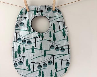 May include: A light blue baby bib featuring a repeating pattern of dark green pine trees and teal ski gondolas on cables. The bib has a rounded neck opening, black snap closures at the top, and a small white fabric tag on the right side edge.