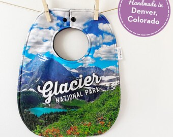 May include: A blue and green baby bib with a picture of Glacier National Park and the text "Glacier National Park". The bib is handmade in Denver, Colorado.