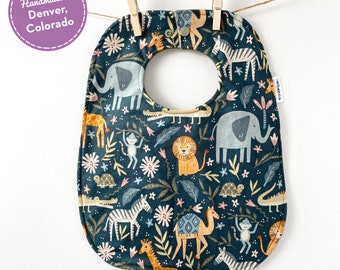 May include: A dark green baby bib with a colorful jungle animal print. The bib features elephants, giraffes, zebras, monkeys, lions, and a camel. The bib is handmade in Denver, Colorado.