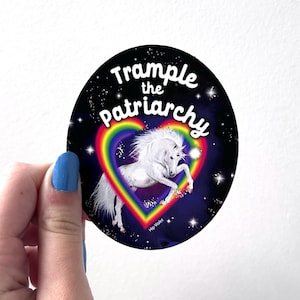 May include: Oval sticker with a white unicorn inside a rainbow heart, set against a starry night sky. The text "Trample the Patriarchy" arches above the heart. The sticker is held by a hand with blue nail polish.