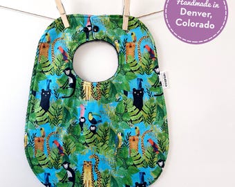 May include: A baby bib with a jungle-themed pattern, featuring monkeys, leopards, parrots, and toucans on a blue background. The bib is held by two wooden clothespins on a string. The text "Handmade in Denver, Colorado" is in a purple circle.