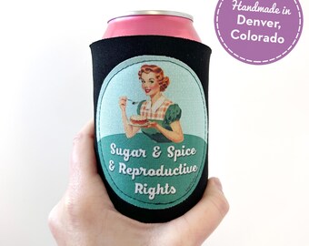 May include: Black can cooler with a retro image of a woman holding a pie, with the text "Sugar & Spice & Reproductive Rights." The can is pink and the cooler is held by a hand. A purple circle reads "Handmade in Denver, Colorado."