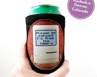 May include: Black can cooler with a retro computer design. The design features a vintage computer with the text "Please be patient I'm from the 1900's". A purple circle with the text "Handmade in Denver, Colorado" is in the background.