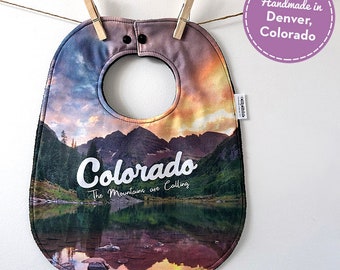 May include: A baby bib with a colorful mountain scene and the text "Colorado The Mountains are Calling".