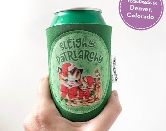 May include: Green can cooler with the text "Sleigh the Patriarchy" and a vintage illustration of two kittens wearing Santa hats and red bows. The cooler is held in a hand.