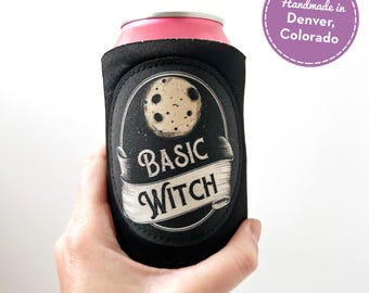 May include: A black foam can cooler featuring a circular patch with a beige moon graphic and the text "BASIC WITCH" printed on a scroll. The cooler is wrapped around a pink aluminum beverage can. A purple circular sticker reads "Handmade in Denver, Colorado".