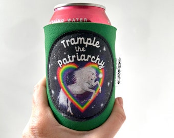 May include: Green can cooler with a black oval patch featuring a white unicorn inside a rainbow heart, set against a starry background. The text "Trample the Patriarchy" is arched above the heart. The can holds a pink can of sparkling water.