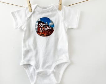 May include: White baby onesie with a round graphic featuring a scenic view of red rock formations under a blue sky. The text "Rock the Cradle" is arched above the image. The onesie is hanging from a clothesline.