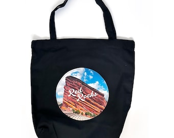 May include: Black canvas tote bag with a white circle featuring a photo of Red Rocks Amphitheatre in Morrison, Colorado. The text "Red Rocks" is printed in white on the photo.