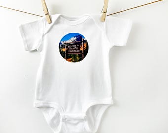 May include: White baby onesie with a round graphic of a mountain scene and a sign that reads "Welcome to Colorful Colorado".
