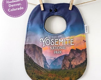 May include: A blue baby bib with a picture of Yosemite National Park. The bib has a black snap closure and a white text overlay that says "Yosemite National Park".