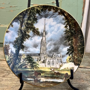 May include: A decorative plate featuring a detailed landscape scene of a cathedral with a tall spire, surrounded by trees and a cloudy sky. The plate has a gold rim and is displayed on a black stand.